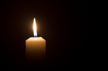 A single burning candle flame or light glowing on a white candle on black or dark background on table in church for Christmas, funeral or memorial service