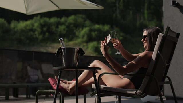 Charming young brunette using modern smartphone for taking selfie while drinking champagne and sunbathing on hotel terrace. Technology, people and vacation concept.