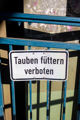 weißes Schild mit der Aufschrift "tauben füttern verboten" in deutscher Sprache an einem blauen Brückengeländer