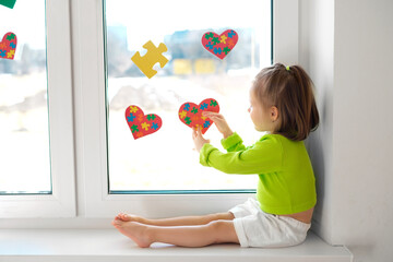 A little girl hangs postcards in the shape of hearts and puzzles on the glass while sitting on the...
