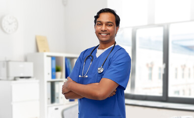 healthcare, profession and medicine concept - happy smiling doctor or male nurse in blue uniform with stethoscope and crossed arms over medical office at hospital background