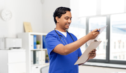 Naklejka na ściany i meble healthcare, profession and medicine concept - happy smiling doctor or male nurse in blue uniform writing medical report on clipboard over medical office at hospital background