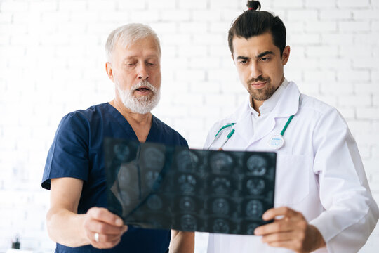 Portrait of senior aged surgeon and young radiologist doctor discussing together history disease of patient using MRI scan of head in hospital office. Concept of modern medicine and health care.