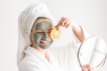 Skin care, beauty, spa. Woman beauty face with mask applying pieces of orange to eyes, in a white bath towel, posing to camera at home, touching face