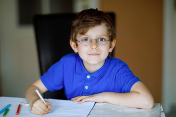 Naklejka na ściany i meble Portrait of little kid boy with glasses at home making homework, writing letters with colorful pens. Little child doing exercise, indoors. Elementary school and education, home schooling concept.