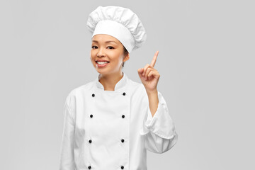cooking, advertisement and food concept - happy smiling female chef in jacket pointing finger up over grey background