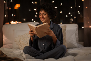 people, bedtime and rest concept - happy smiling woman in pajamas reading book sitting in bed at...