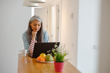Female person standing by wooden table with cup of coffee and using modern laptop in kitchen