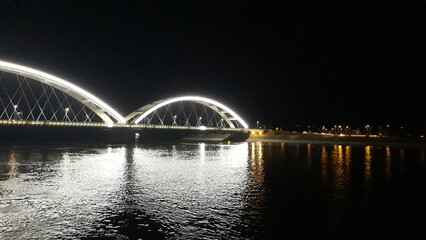 Fototapeta na wymiar Die Brücke über den Fluss wirft Weiß in die Nacht. Die Lichter der nächtlichen Stadt spiegeln sich im Fluss. Silhouetten von Siedlungen am See. Verschwommene Fotos in der Dunkelheit der Nacht.