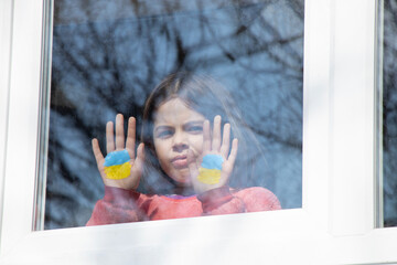 A little girl in the window of her house with a painted yellow-blue flag of Ukraine, peace in...
