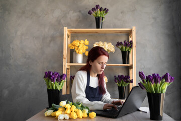 Portrait florist in a blue work apron working in her own flower shop, using a laptop, organizing...