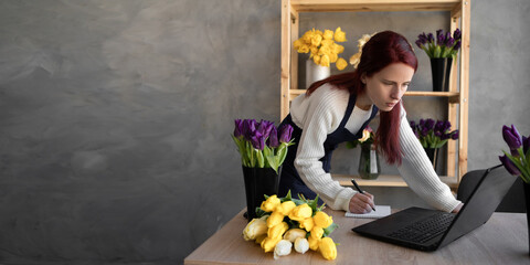 Small business. a female florist in a flower shop near a showcase with tulips in an apron takes an order online using her laptop. Banner. - Powered by Adobe