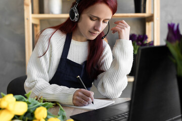 Young beautiful female florist in blue apron business owner using headset and computer while talking to customers online taking order online.