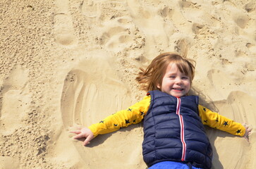 Little boy, 3 years old, lies in the sand on the beach and makes a "snow angel". Child laughing on the beach