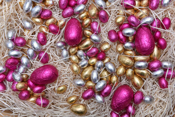 Pile of colorful pale pastel foil wrapped chocolate easter eggs in pink,  silver and gold, on pale shreaded cream nest.