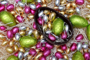 Pile of colorful foil wrapped chocolate easter eggs in green, pink, yellow and gold with half of a large brown dark chocolate egg in the middle and mini eggs inside, on pale shreaded cream nest.