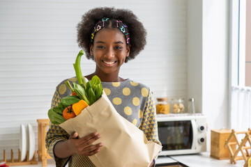 Young beautiful African black girl woman in kitchen holding paper grocery shopping bag full of...