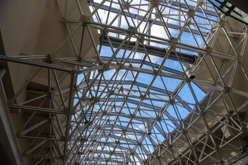 Modern architectural fiction. A metal structure supporting contemporary gallery glass dome. Transparent ceiling letting in sunlight, glass dome blue sky and rooftop in background.