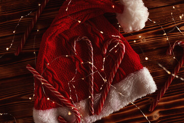 Candy canes on a red Christmas hat among garlands on a wooden table top view