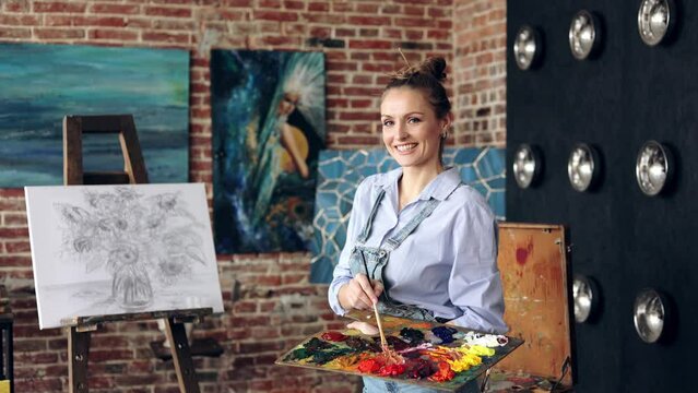Portrait of female artist mixing color paints with art brush on the palette. Young beautiful painter at the studio preparing for painting a still life picture with sunflowers.