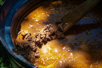 Mixing and cooking pork and beef goulash (stew) with wooden mixing spoon in big caldron (aluminium pot). Traditional cooking on campfire. Close-up.