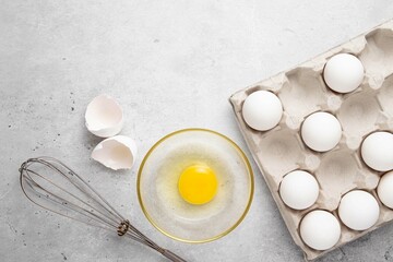 Fresh white organic chicken eggs in paper tray, broken egg and shell, whisk on light background. Baking background. Top view, copy space or empty place for text.