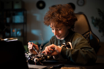 A smiling boy is learning electronics and soldering wires and circuit boards in the field of robotics. A child works on a toy in room late at night.