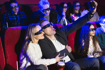 Beautiful girl and guy laughing happily making a selfie together during a movie at the cinema. Young couple watching a movie at a cinema and photographing themselves with a smartphone. 