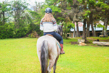 Back view of a caucasian woman wearing a andean sweater riding a horse near a forest.