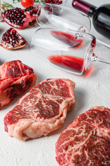 Raw set of alternative beef cuts Chuck eye roll, top blade, rump steak with red wine in glass and bottle, herbs and pomegranate. Organic meat. White textured background. Side view vertical.