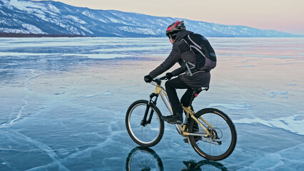 Man is riding bicycle on the ice. Ice of the frozen Lake Baikal. Teenage is dressed in black down jacket, cycling backpack, helmet. Tires on covered with special spikes. Traveler boy is ride cycle.