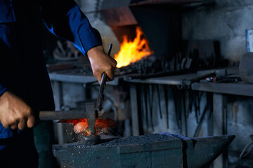 Close up worker in blue uniform keeping hammer and creating nice decorations for fence in large modern smithy. Concept of process creating decorations in professional workshop.