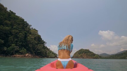 Sexy young woman in blue swimsuit swings feet in water on pink canoe on sea against hills backside view. Traveling to tropical countries. Attractive sportive girl is sailing on kayak in ocean.