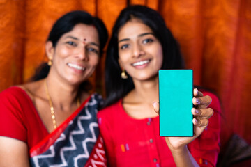 Indian mother and daughter showing green screen mobile phone by looking at camera -concept of...