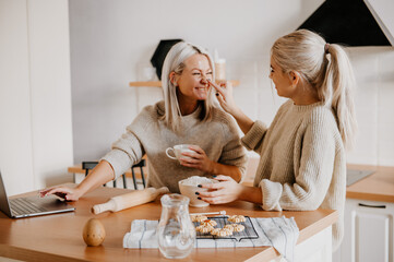 middle aged blonde mother and teen daughter in kitchen watching recipe or tv show on laptop and cooking. lifestyle natural concept