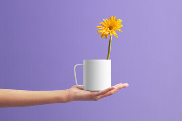 White cup of tea stands on the hand at the violet background. Yellow flower in the white cup.