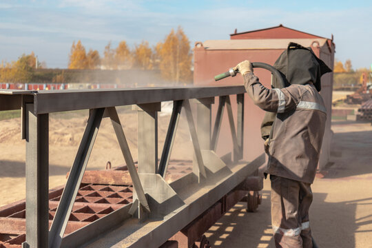 A worker in special clothing is sandblasting a metal building structure at an industrial site. Industrial metal processing.