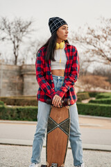 Beautiful young hipster woman posing with skateboard. Hispanic Latin woman wearing casual clothes.