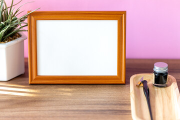 Blank landscape wooden frame beside a small houseplant and calligraphy tools, painting or artwork display mockup in front of pink wall
