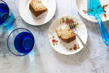 Two slices of banana cakes on white plate with fork and blue glasses of water on concrete table top in top view.
