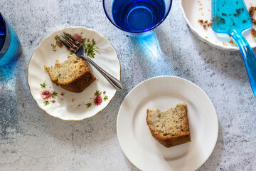 Two slices of banana cakes on white plate with fork and blue glasses of water on concrete table top in top view.