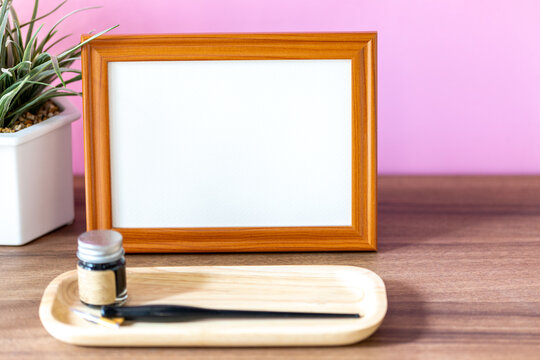 Blank landscape wooden frame beside a small houseplant and calligraphy tools, painting or artwork display mockup in front of pink wall