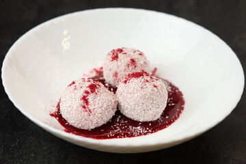 Homemade coconut balls, raw and healthy sugar free rafaello candies. Vegetarian energy sweets recipe. Round tasty dessert in bowl with strawberry sauce on dark background. Copy space, close up view.