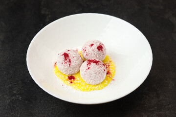 Homemade coconut balls, raw and healthy sugar free rafaello candies. Vegetarian energy sweets recipe. Round tasty dessert in bowl with yellow mango sauce on dark background. Copy space, close up view.
