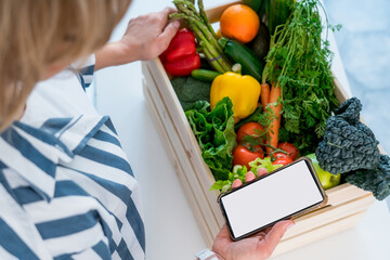 Top view woman holding wooden box with fresh vegetables and phone with blank white screen. Mockup...