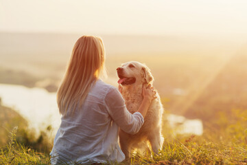 Blonde woman enjoying nature and embracing pet dog on the hill at sunset. 