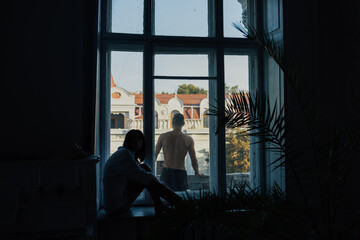 Fototapeta na wymiar Romantic young couple relax at home. Man is standing on the on the balcony with cityscape and woman sitting on the windowsill.