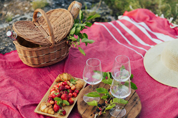 Fototapeta na wymiar Picnic with glasses of white wine on a rock. Three glasses of white wine, berries, croissants, hat and basket on red cover.