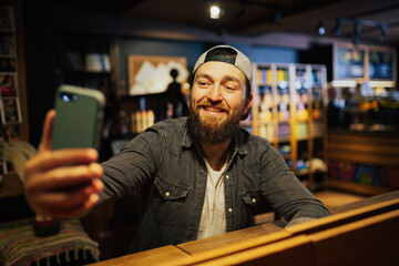 Handsome happy bearded man is doing selfie using a smartphone and smiling while sitting in the bar counter in pub.