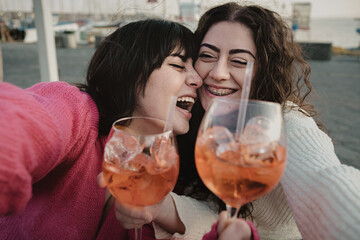 Cheerful best friends selfie with girls drinking spritz cocktail at the outdoors pub cafe - happy young women having fun
